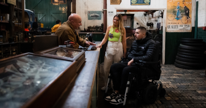 Woman stands next to man in wheelchair as they talk to a staff member inside Beamish Museum garage exhibition at the 1900s Town.